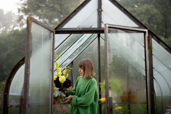 Eine Frau in einem grünen Pullover steht im Gewächshaus und hält einen Blumenstrauß mit Sonnenblumen und anderen Blumen.